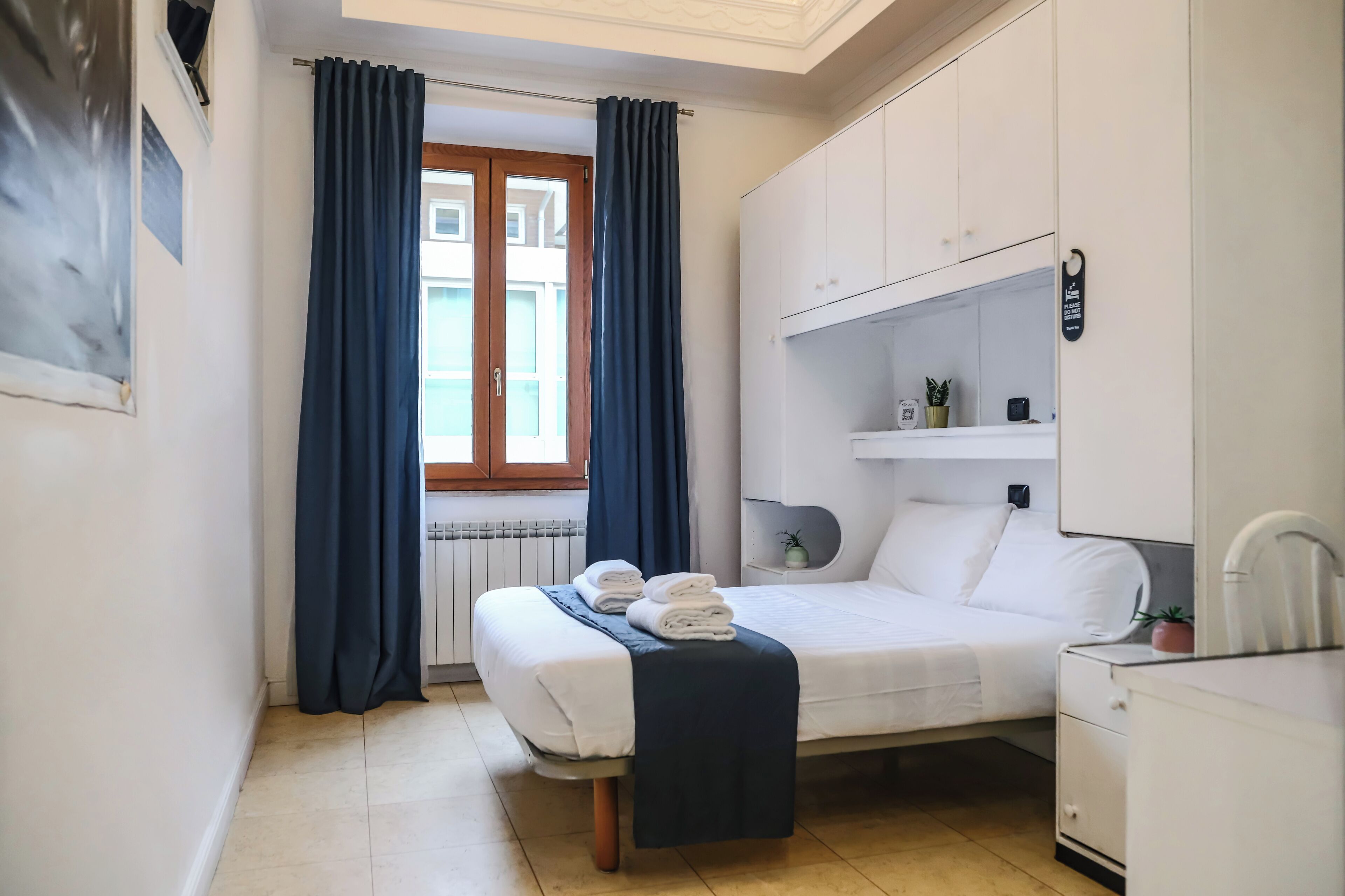 A small, neatly arranged bedroom with a bed, bedside table, and a window with curtains.