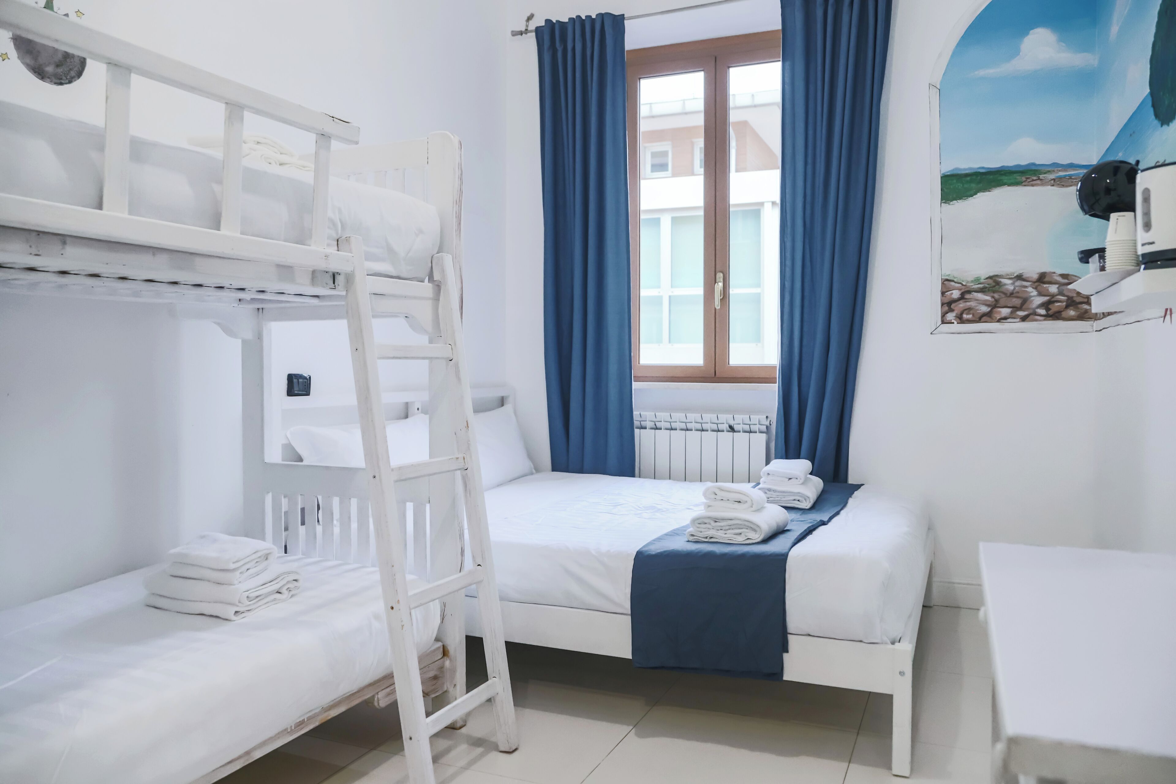 Habitación con literas, una ventana, una cama con manta azul y una mesa blanca.
