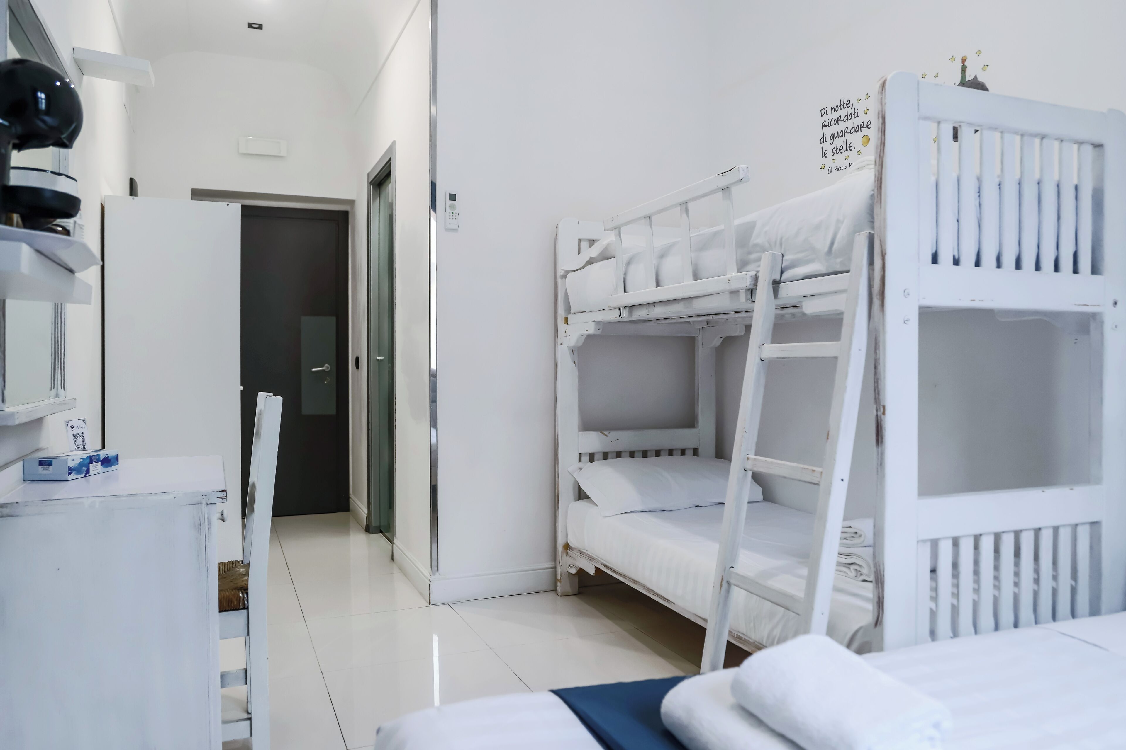 Une pièce avec des lits superposés, un bureau avec une chaise, une petite table sur laquelle sont posés divers objets, et une porte qui donne sur une autre pièce.