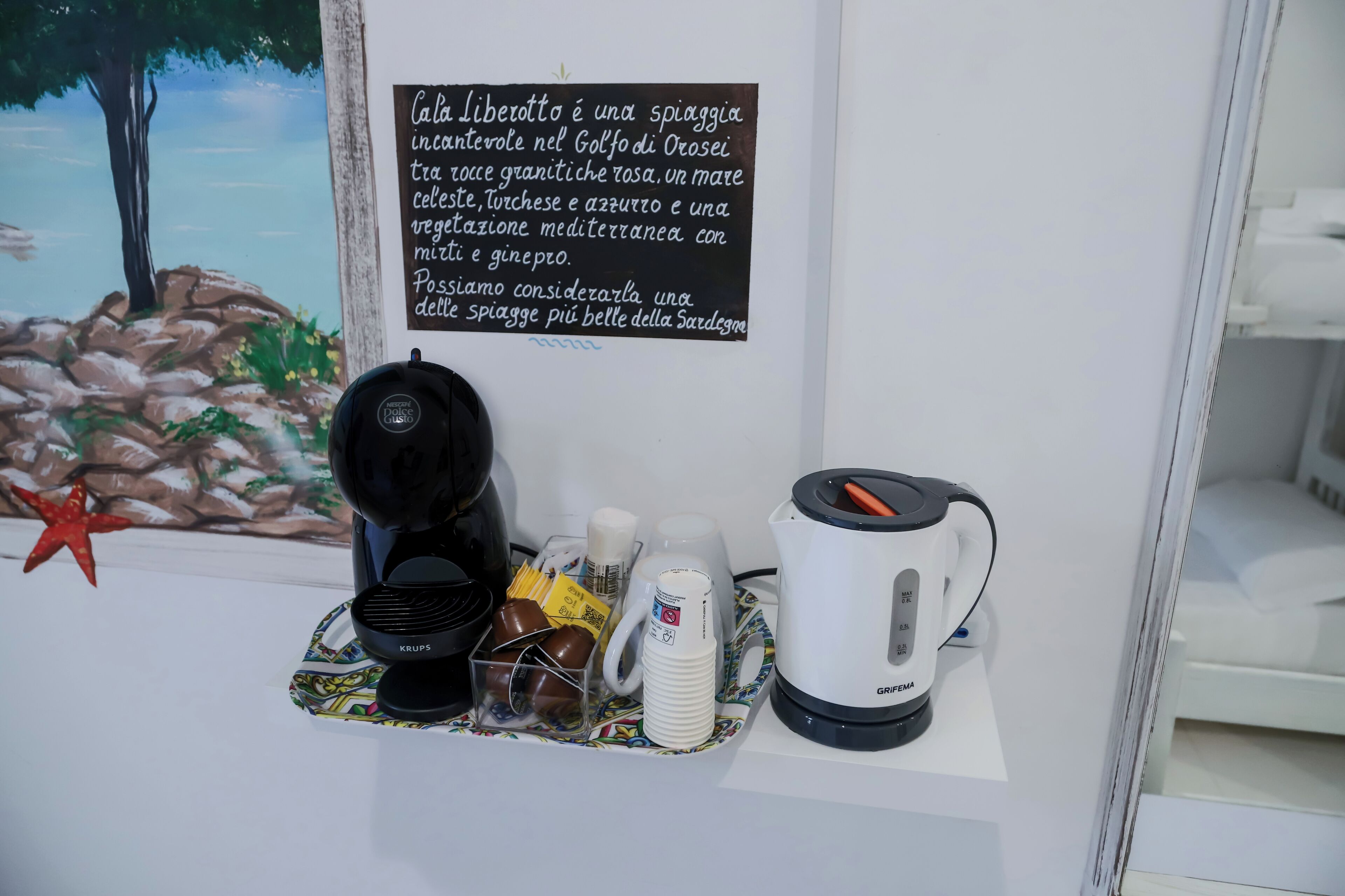 Een koffiehoek met een Krups koffiezetapparaat, een witte koffiekan en een bordje met informatie over Cala Liberetto.