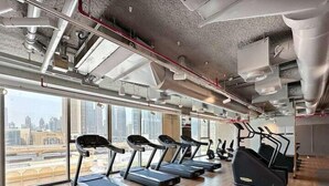 Ein Fitnessstudio mit Laufbändern und Trainingsgeräten, großen Fenstern mit Stadtblick und freiliegenden Lüftungsrohren an der Decke.