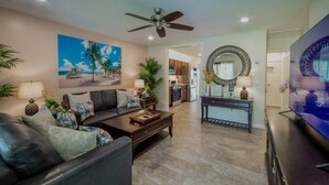 A living room with a dark leather sofa, a coffee table, and a TV. There's a painting of a beach scene on the wall, a ceiling fan, and a kitchen in the background. - 2 King Beds, Kitchen, Living & Dining Room, Yard, Heated Pool, WiFi, Apt#2112 (Wilton Manors)