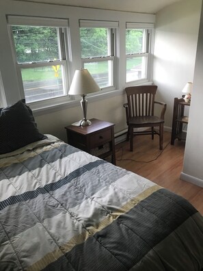 Un dormitorio con cama, una silla, una mesita de noche, una lámpara y una ventana con vistas a árboles.