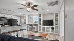 A modern kitchen with a microwave, refrigerator, and bar stools. There is a TV on a cabinet and a ceiling fan above the kitchen. - Cozy Cottage w/ Gorgeous Views - Near the beach! (Galveston)
