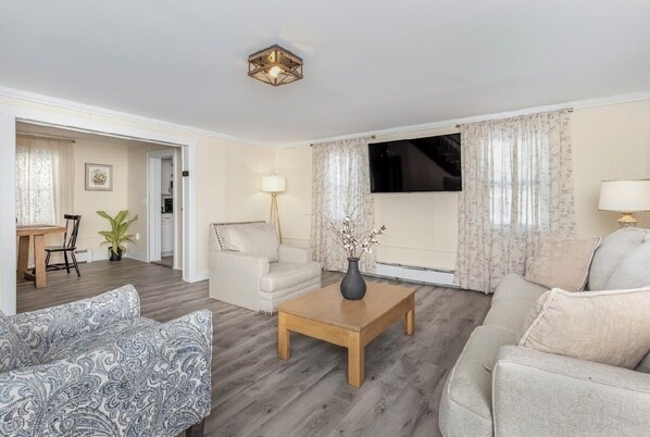 Un salón con sofá, sillón y otro sillón estampado, una mesa de centro de madera con un jarrón, y un televisor montado en la pared.