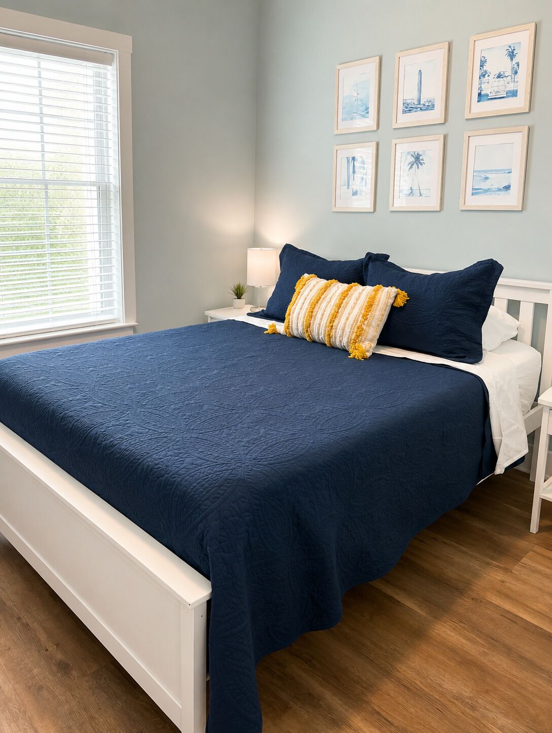 Un dormitorio con cama, mesitas de noche blancas, una lámpara y cuadros en la pared.