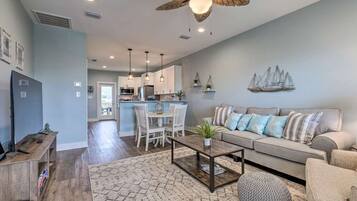 A modern living room with a sofa, coffee table, and a ceiling fan. There is a kitchen area in the background with a dining table and chairs.