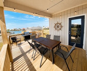 A covered outdoor deck with a wooden table and chairs, a grill, and a view of the water.