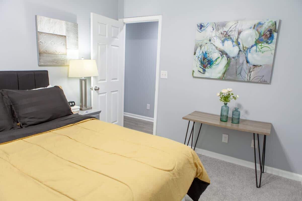 A bedroom with a bed, a nightstand, a lamp, a painting, and a small table with vases.