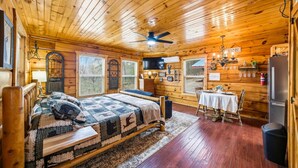 A cozy cabin with a wooden interior, a bed with a quilt, a dining table with chairs, and a ceiling fan. - Romantic Hot Tub Cabin w/ Fast WiFi | Near Smokies (Sevierville)