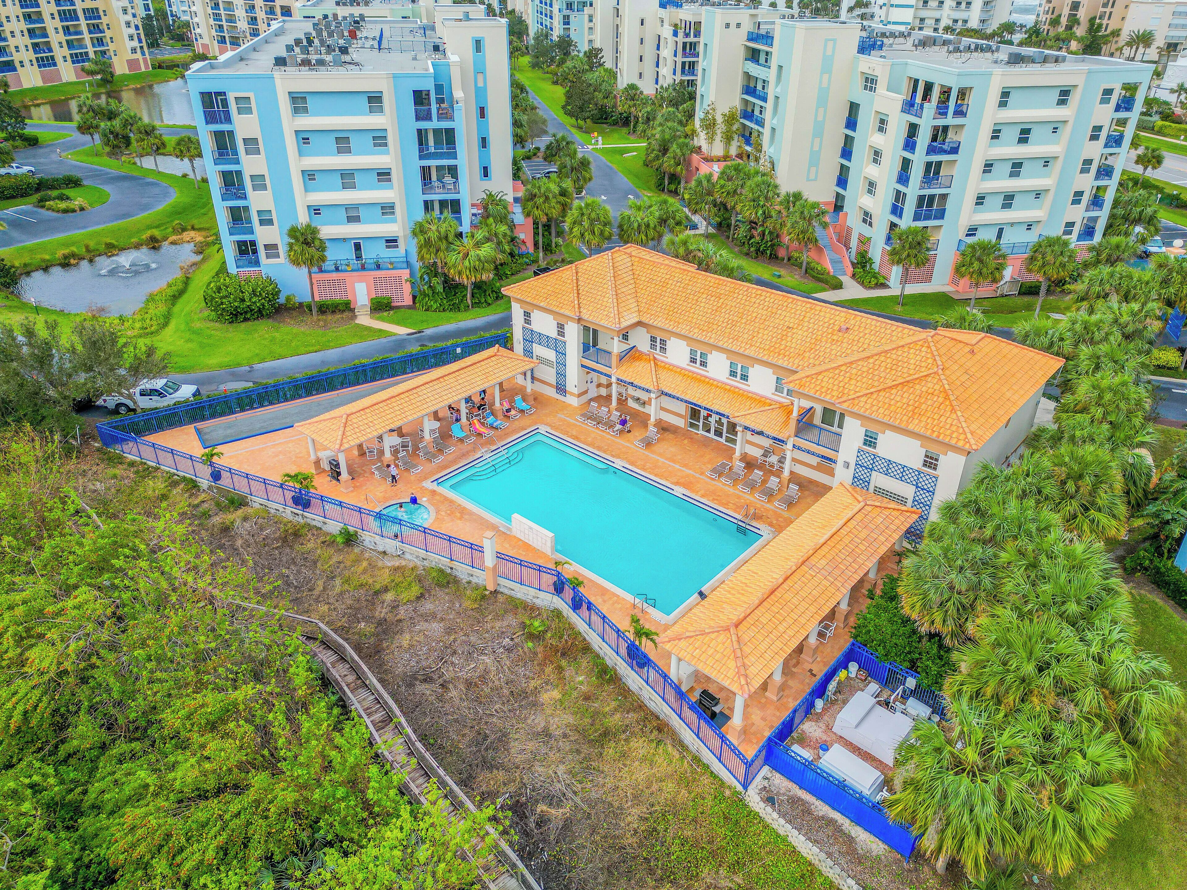 A large building with a swimming pool surrounded by a blue fence, multiple balconies, and lush greenery.