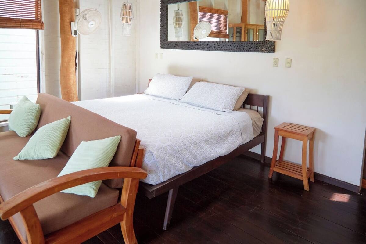Une chambre à coucher avec un lit, une chaise en bois, un petit tabouret en bois et un ventilateur fix é au mur.