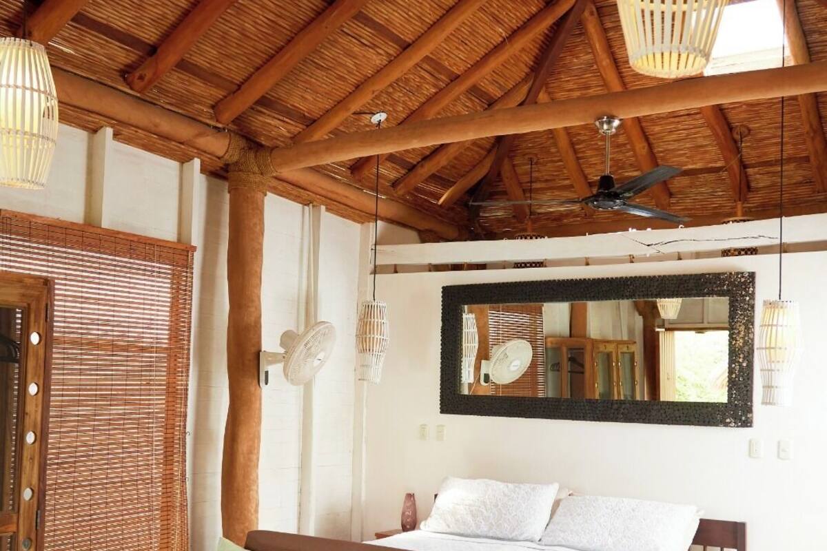 Une pièce spacieuse avec un haut plafond en bois, un grand lit recouvert de draps blancs, un canapé en bois agrémenté de coussins verts et une table en bois sur laquelle est posé un vase.