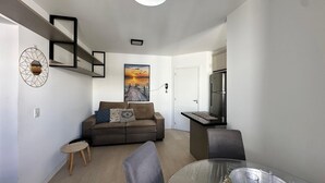 A modern living room with a sofa, a coffee table, and a dining area with chairs. There is a refrigerator and a painting on the wall. - PCM402VG21 (Florianópolis)