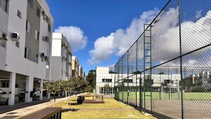 A courtyard with a wooden bench, a tennis court enclosed by a fence, and modern buildings in the background. - PCM402VG21 (Florianópolis)