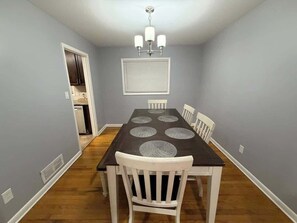 Zona pranzo con tavolo in legno scuro, sedie bianche, lampadario a soffitto e bocchetta di aerazione sulla parete.