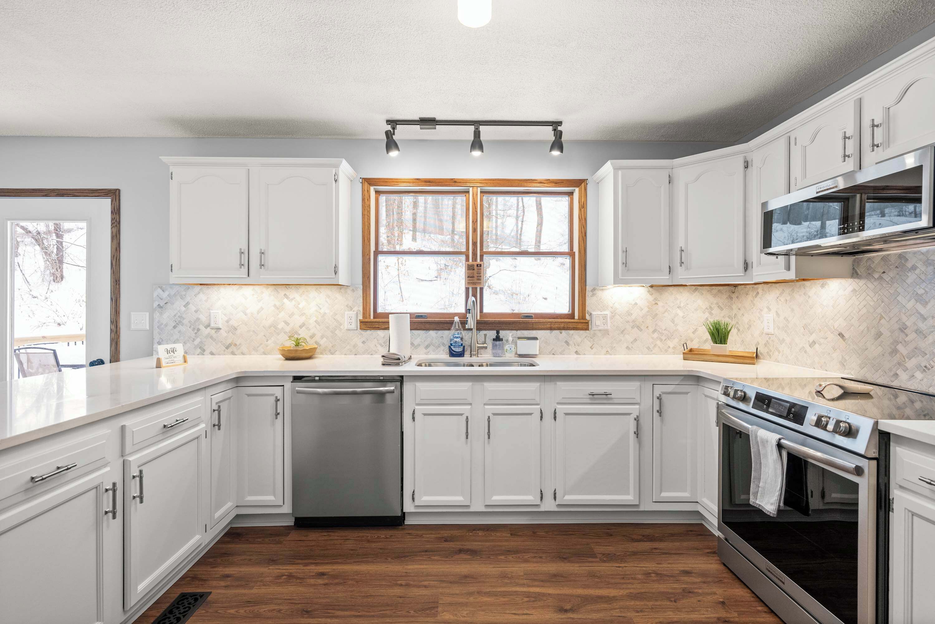 A modern kitchen with white cabinets, a stainless steel dishwasher, an oven, and a window with a view of trees.
