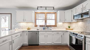 A modern kitchen with white cabinets, a stainless steel dishwasher, an oven, and a window with a view of trees.
