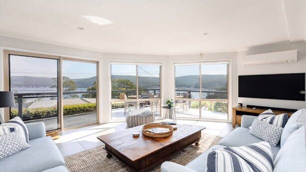 A spacious living room with large windows, a flat-screen TV, and a view of a lake and hills. - Bay views with a secluded bush backdrop (HARDYS BAY)
