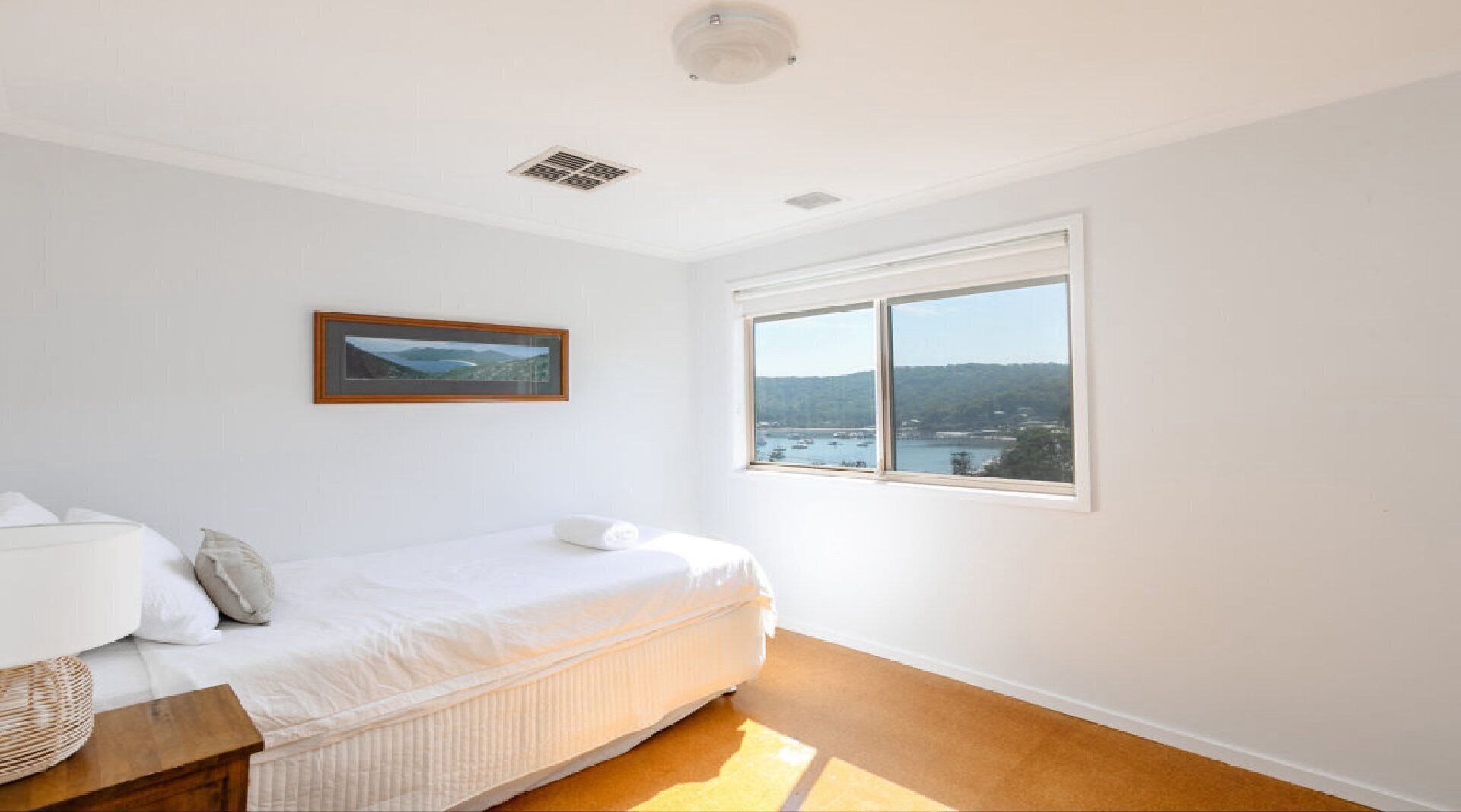 A bedroom with a bed, a window with a view of a body of water and hills, and a ceiling vent.