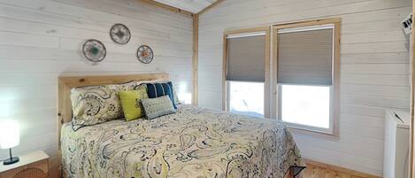 A bedroom with a wooden floor, a bed with patterned bedding, a nightstand, a ceiling fan, and a window with blinds.
