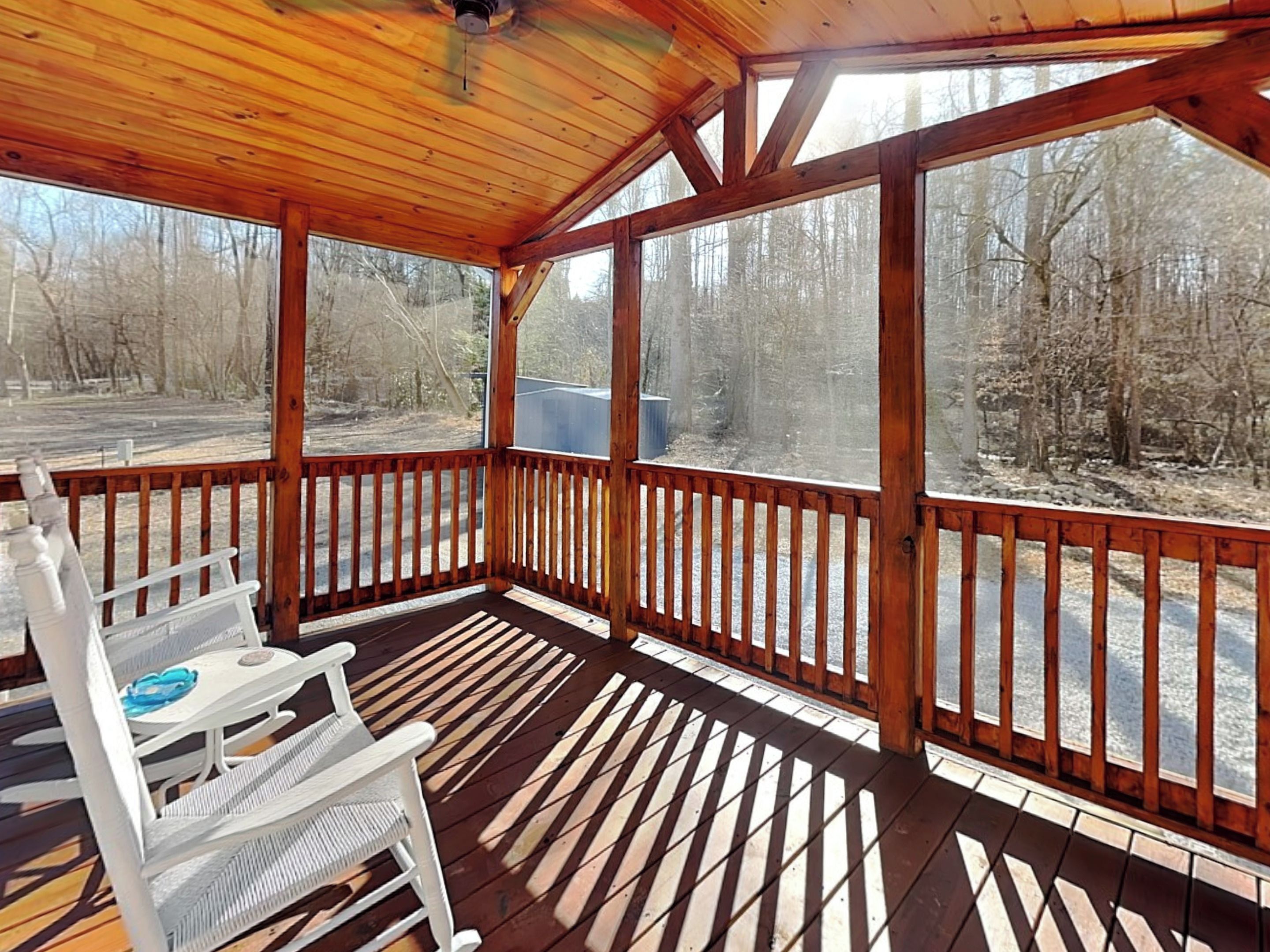 A covered outdoor area with a wooden deck, white metal chairs, and a view of a forested area.