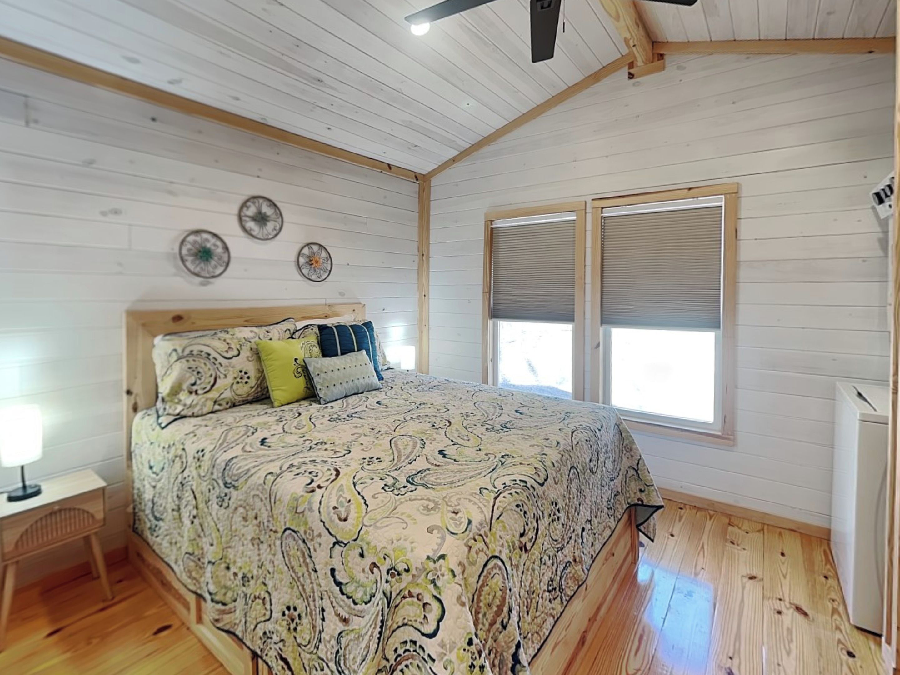 A bedroom with a wooden floor, a bed with patterned bedding, a nightstand, a ceiling fan, and a window with blinds.