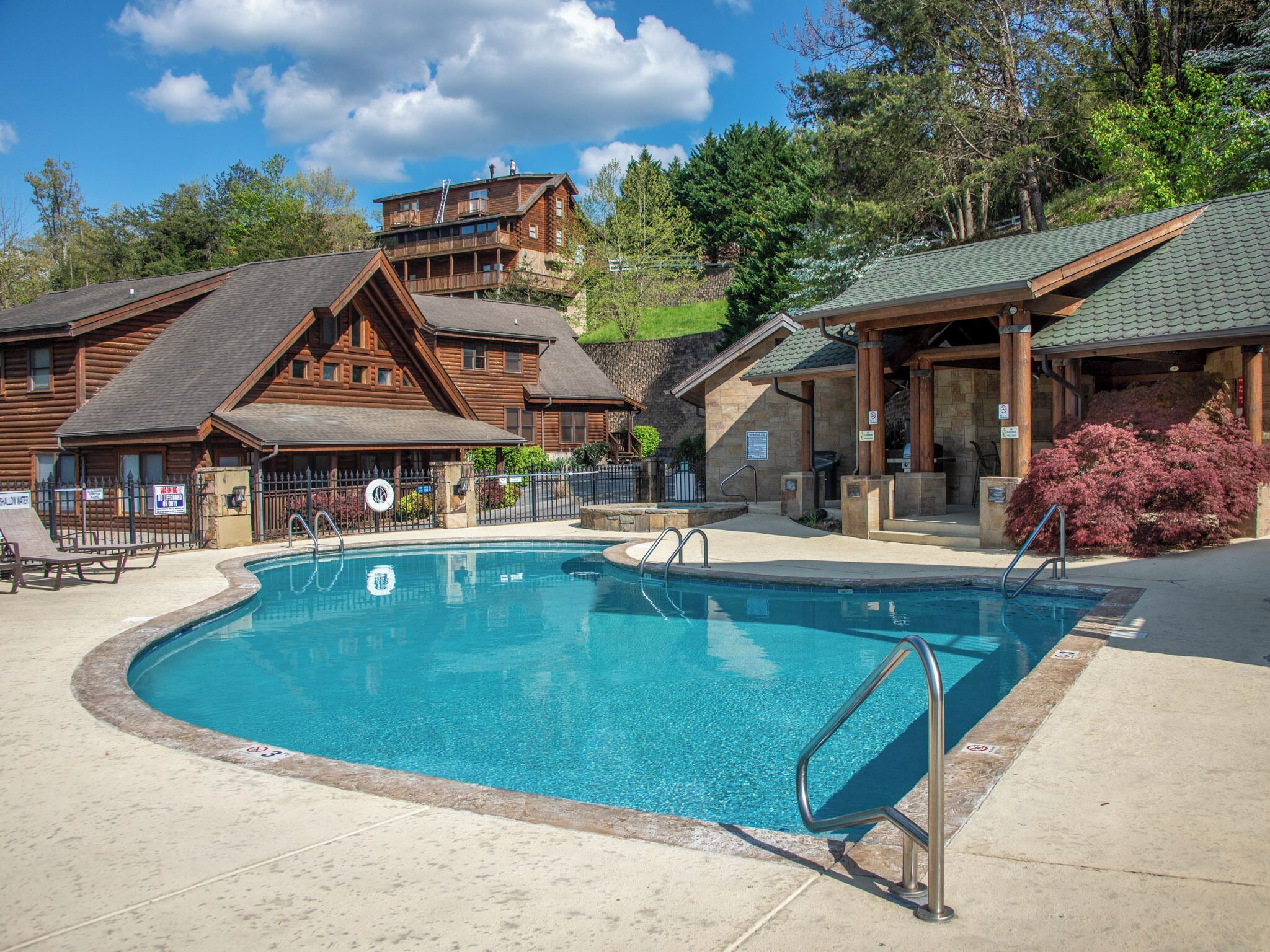 A pool area with a wooden lodge in the background, a covered seating area, and a sign indicating shallow water.