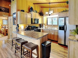 A kitchen with wooden cabinets, a granite countertop, a stainless steel refrigerator, and a central island with bar stools. - The Grand View (Gatlinburg)