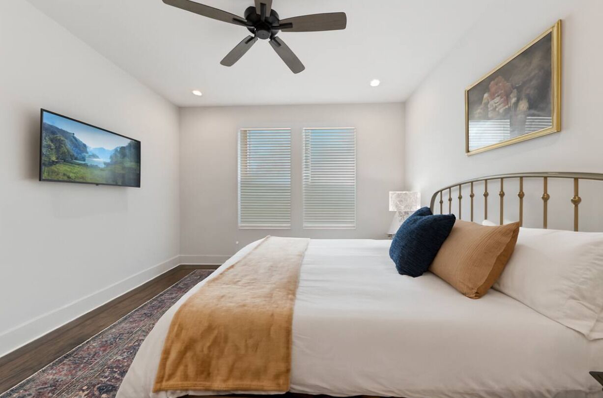 Un dormitorio con cama, ventilador de techo, un cuadro y ventana con persianas.