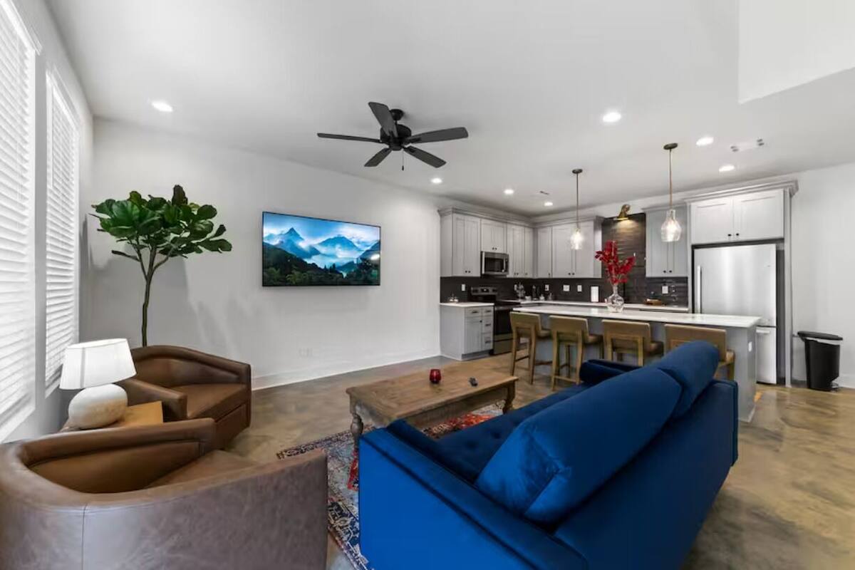 A modern living room with a blue sofa, a wooden coffee table, and a brown leather chair. A ceiling fan is visible, and there is a kitchen area with white cabinets and a refrigerator in the background.