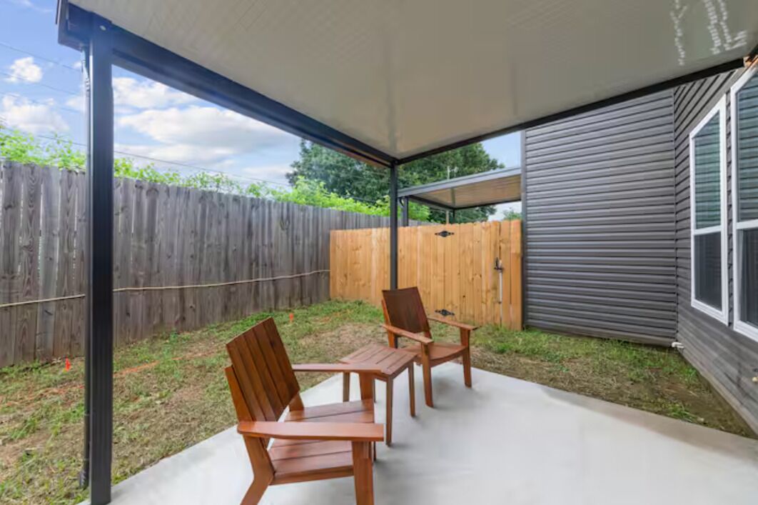 A covered patio with two wooden chairs and a table, a wooden fence in the background, and a house with a grey exterior.