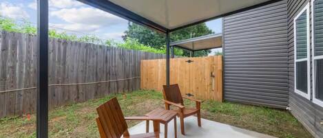 A covered patio with two wooden chairs and a table, a wooden fence in the background, and a house with a grey exterior.