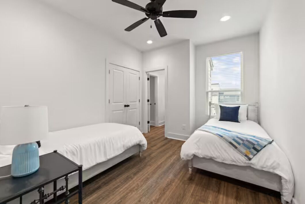 A bedroom with two single beds, a ceiling fan, a window with blinds, and a nightstand with a lamp.