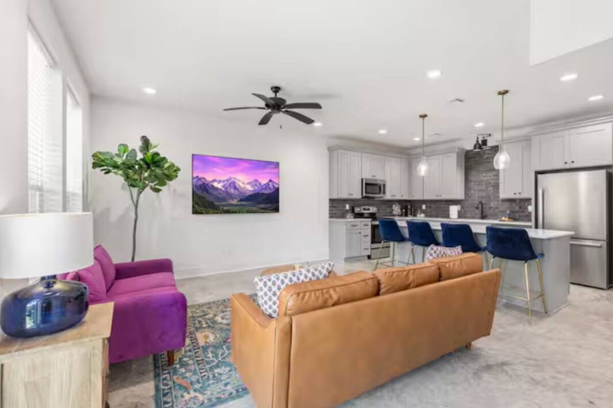 A modern living room with a purple sofa, a brown sofa, and a coffee table. There is a kitchen area with white cabinets, a refrigerator, and a dining table with blue chairs.