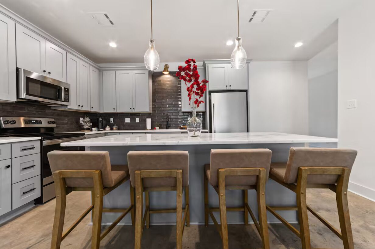 Una cocina moderna con armarios blancos, una isla central con taburetes de bar, electrodomésticos de acero inoxidable e iluminación colgante.