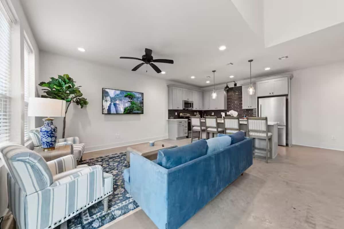 A spacious living room with a blue sofa, a striped armchair, and a coffee table. There is a kitchen area with white cabinets and a dining table.