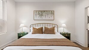 A neatly made bed with a headboard, two bedside tables with lamps, a wall-mounted artwork, and a window with blinds.