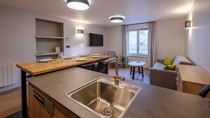Une cuisine moderne dotée d’un évier en acier inoxydable, d’un plan de travail en bois et d’étagères intégrées. On y trouve un coin repas avec une table et des chaises, ainsi qu’un canapé dans l’espace salon.
