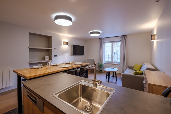 Une cuisine moderne dotée d’un évier en acier inoxydable, d’un plan de travail en bois et d’étagères intégrées. On y trouve un coin repas avec une table et des chaises, ainsi qu’un canapé dans l’espace salon.