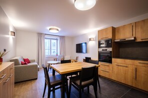 A modern kitchen with wooden cabinets, a dining table with chairs, a sofa, and a TV mounted on the wall.