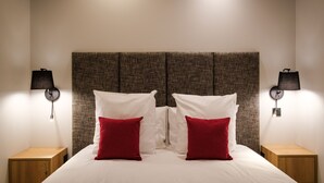 Un lit bien fait, avec une literie blanche et deux oreillers rouges, de part et d’autre duquel se trouvent deux tables de chevet avec des lampes.