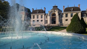 Une grande fontaine se trouve devant un bâtiment historique, doté de nombreuses cheminées et d’une entrée centrale en arc de cercle.