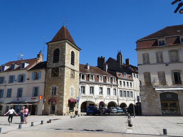 A historic building with a clock tower, multiple arches, and a paved square with parked cars.