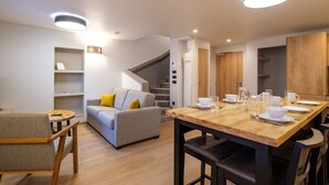 A modern living room with a grey sofa, wooden coffee table, and a dining area with a wooden table set for four. - Grand T3 familial en centre-ville (Lons-le-Saunier)