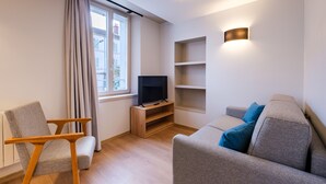 Un salon moderne avec un canapé gris, une chaise en bois, un téléviseur posé sur un meuble en bois et une fenêtre avec des rideaux.
