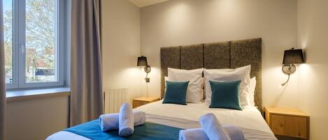 A neatly made bed with white and teal bedding, two bedside lamps, and a window with curtains.