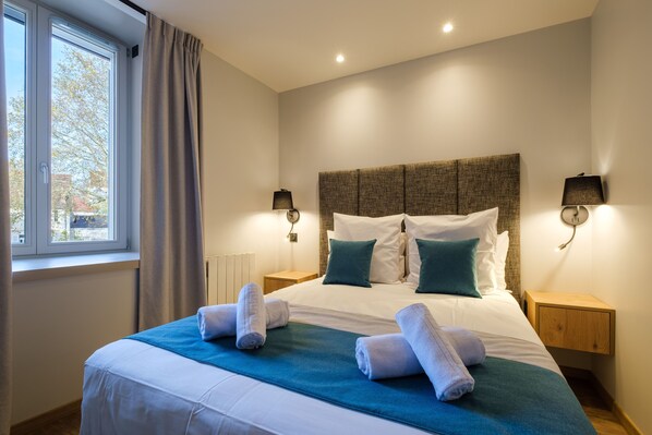 Una cama bien tendida con ropa de cama blanca y verde azulada, dos lámparas de noche y una ventana con cortinas.
