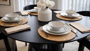 Una mesa redonda de comedor con platos, cuencos y cubiertos blancos, adornada con individuales tejidos y un jarrón con flores blancas.