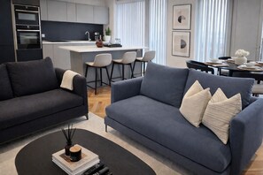 Un salon moderne avec un canapé bleu foncé, une table basse avec des livres et une bougie, et une cuisine en arrière-plan.
